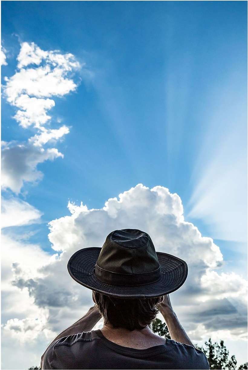 Person wearing a hat, looking at clouds in the sky with sunbeams shining through, creating a beautiful nature scene.