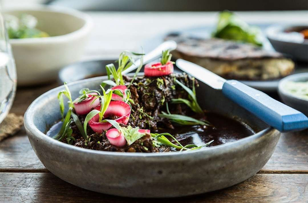 Close-up of dark saucy dish in ceramic bowl with garnish and knife on a rustic wood table. Food presentation for restaurant menu.