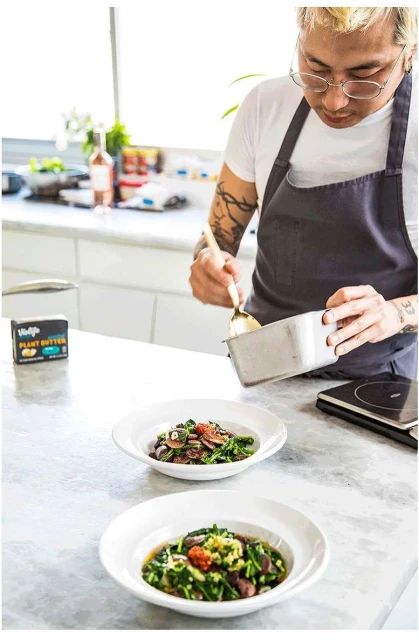 Chef plates a vibrant vegan dish in a bright kitchen. Fresh ingredients, plant-based butter, and stainless steel utensils complete the culinary scene.