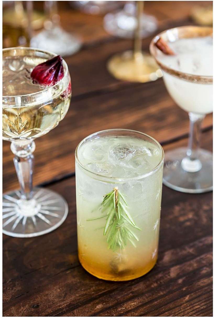 A cocktail in a clear glass with a sprig of rosemary on a wood table, surrounded by other stemmed cocktails and glassware.
