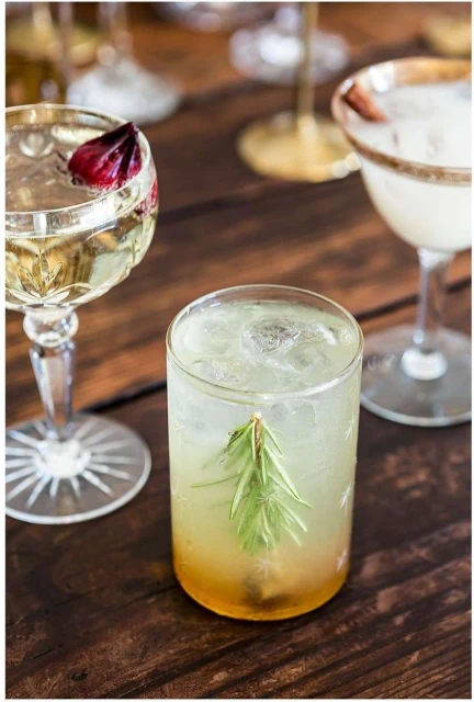 A cocktail in a clear glass with a sprig of rosemary on a wood table, surrounded by other stemmed cocktails and glassware.