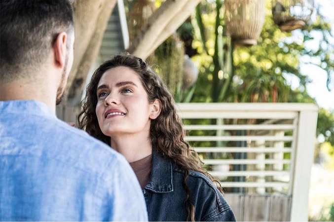 Close-up portrait of a smiling woman with long brown wavy hair looking up at a man in a blue shirt outdoors.
