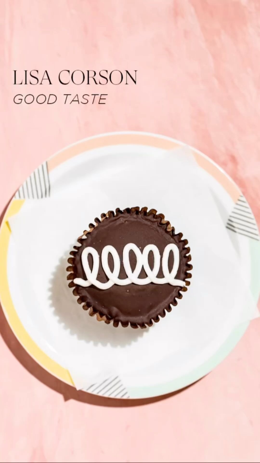 Close-up of a single chocolate cupcake with white frosting swirls, displayed on a patterned plate against a pink backdrop.
