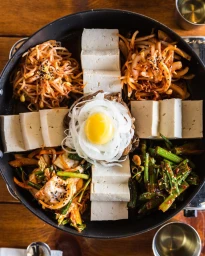 Overhead shot of a large round skillet containing neatly arranged tofu blocks, seasoned bean sprouts, sautéed vegetables, and a fried egg.