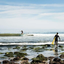 Surfer in a black wetsuit walking into the ocean carrying a yellow surfboard, with a second surfer riding a wave in the distance.