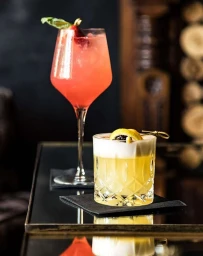 Two cocktail drinks on a dark glass table, a tall pink spritz and a short yellow sour with a lemon twist
