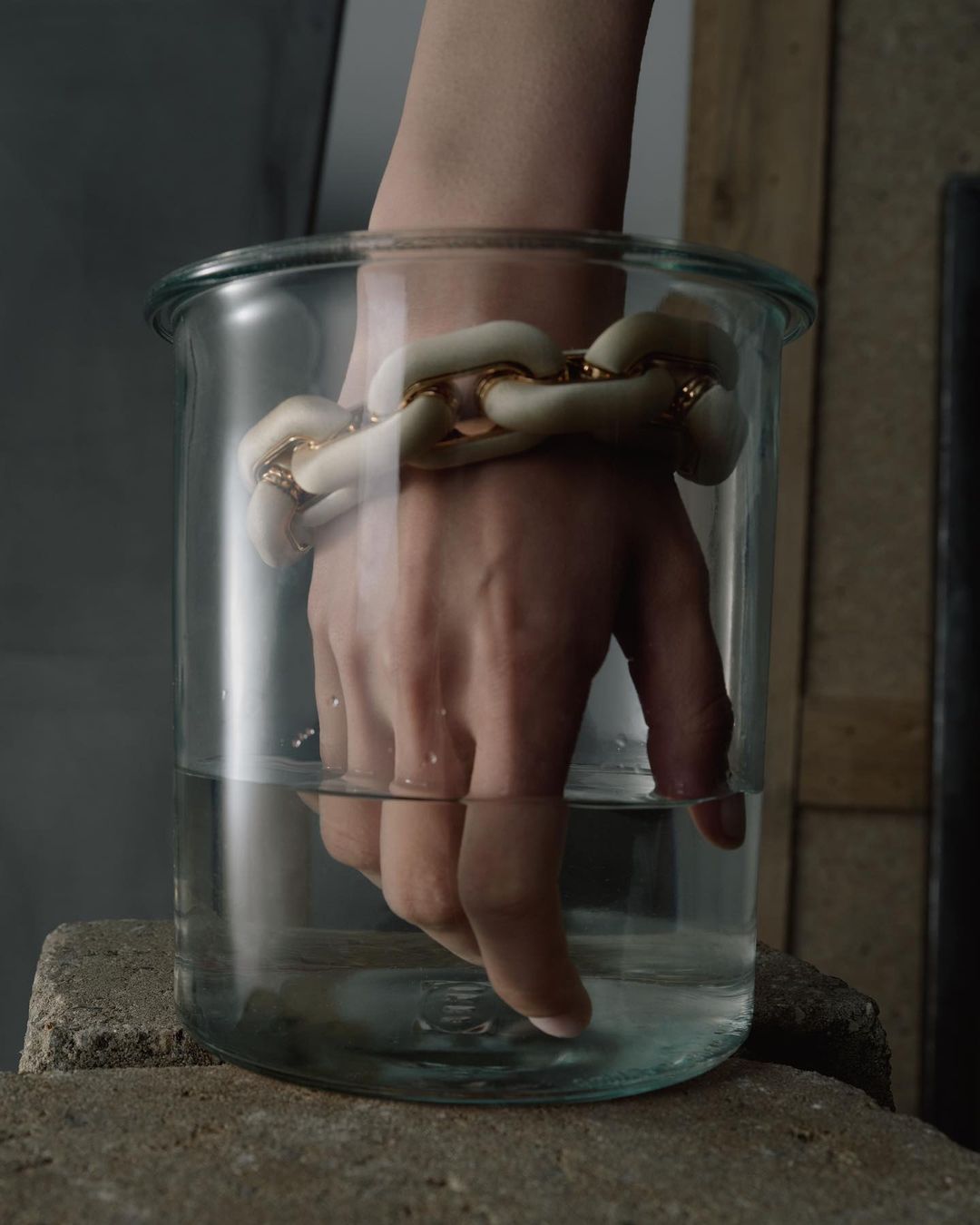 Hand submerged in a glass vase filled with water, wearing a chunky chain bracelet with white and gold links, minimalist background.