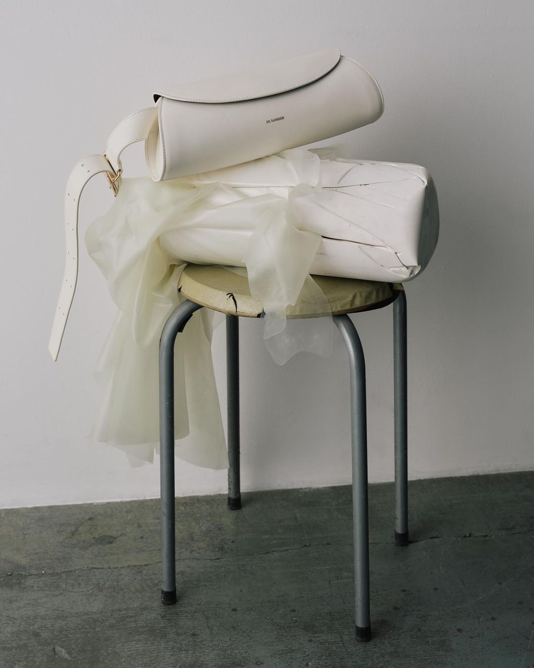 Three white handbags and a sheer textile sit on a metal stool in a studio setting. The minimalist product shot features a neutral color palette.