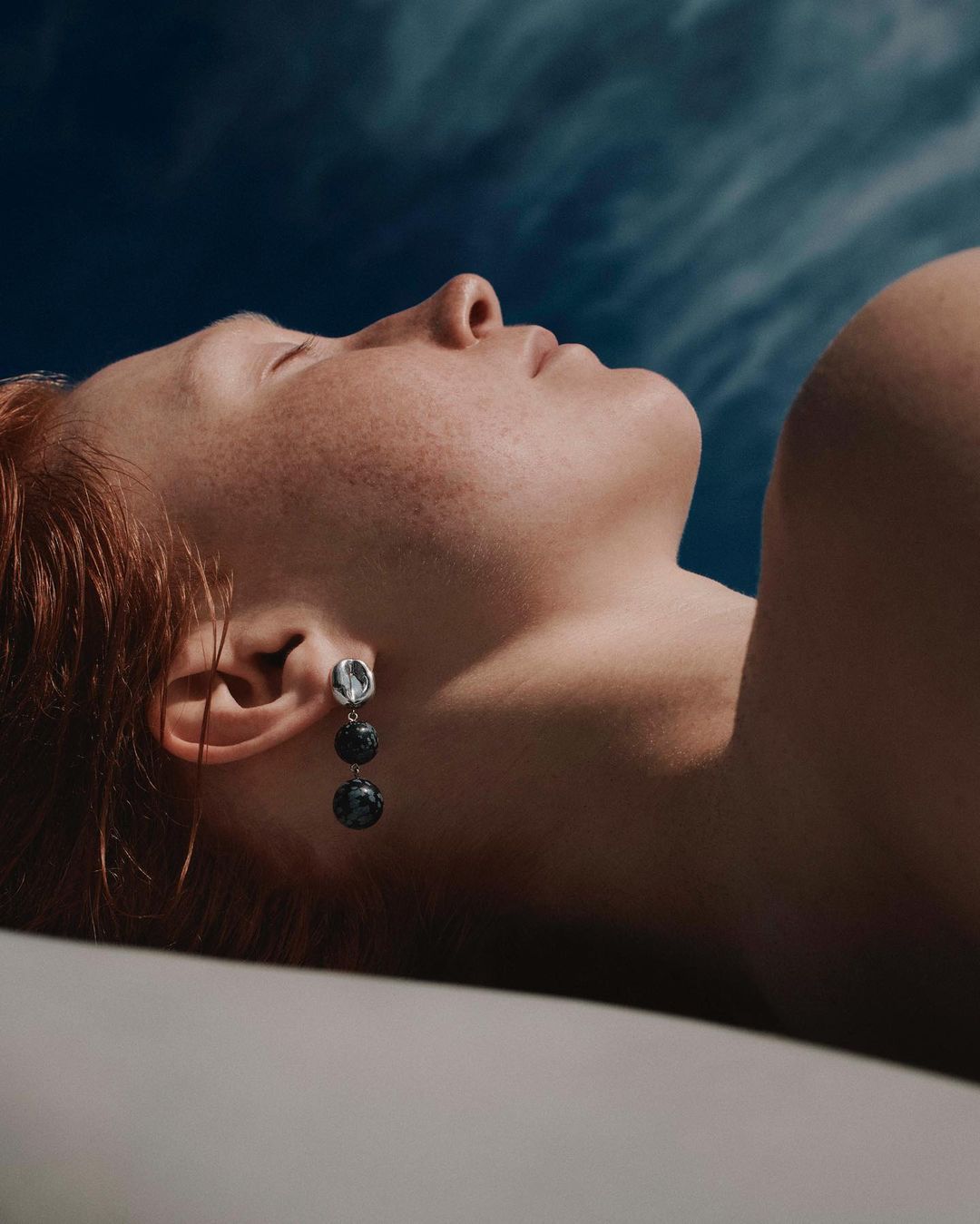 Woman with red hair and an earring with black pearls, laying in white sheets against a blue sky, fashion, beauty, portrait photography.