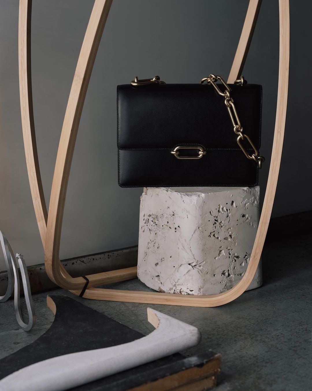 Black leather handbag with a gold chain strap is displayed on a concrete block with a wood structure in the background.