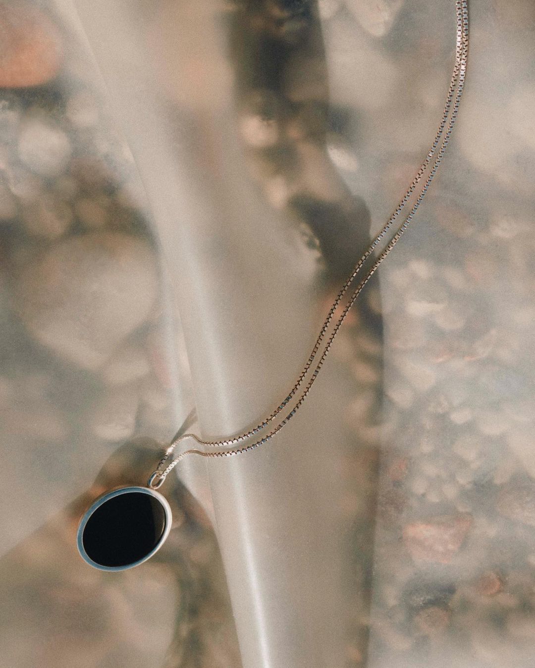 Close-up of a silver chain necklace with an oval black pendant against an abstract background with beige and white tones.