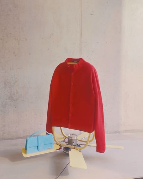 Still life of a red sweater and a light blue purse sitting on top of a vintage-style fan in front of a concrete wall.
