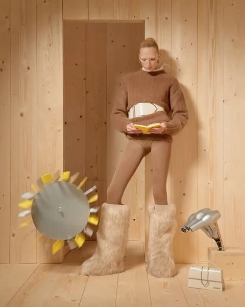 Blonde woman in furry boots and brown sweater reads a book in a wood-paneled room with a toy turbine and hairdryer.
