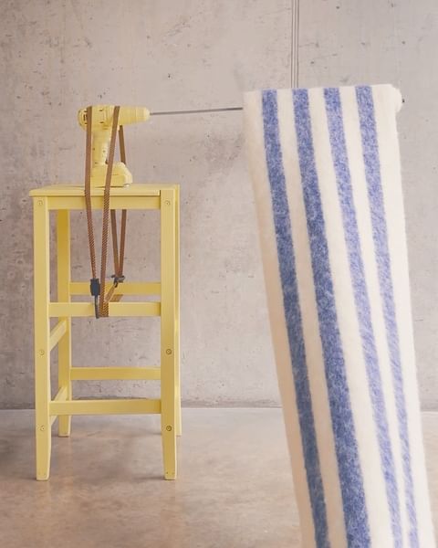 Still life featuring a yellow painted stool with a yellow power drill and a blue and white striped fabric against a concrete wall.