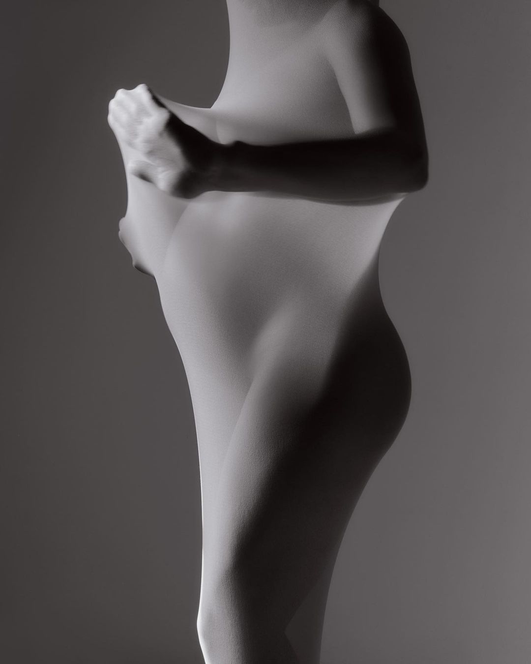 Figure of a woman wrapped in white fabric, with hands visible pressing the fabric against her form, in a monochrome portrait.