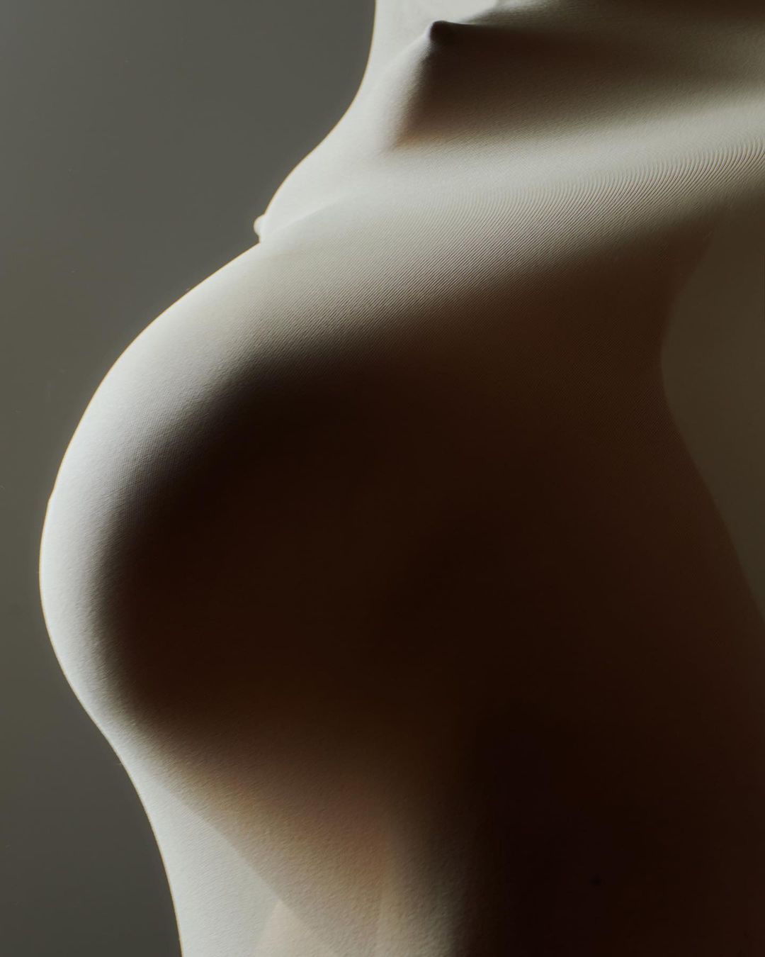 A side view of a pregnant adult female figure covered with light fabric, showcasing the form with soft lighting in a studio setting.