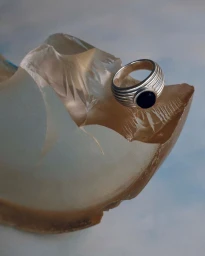 Silver ring with a dark stone resting on a translucent amber glass sculptural form against a pale blue background.