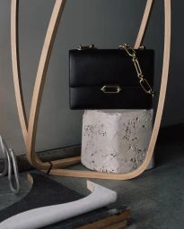 A black leather handbag with a chunky gold chain strap resting on a textured white block inside a sculptural wooden frame.
