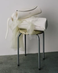 White leather cylindrical handbags stacked on a stool with sheer draped fabric against a plain wall by Lisa Jahovic