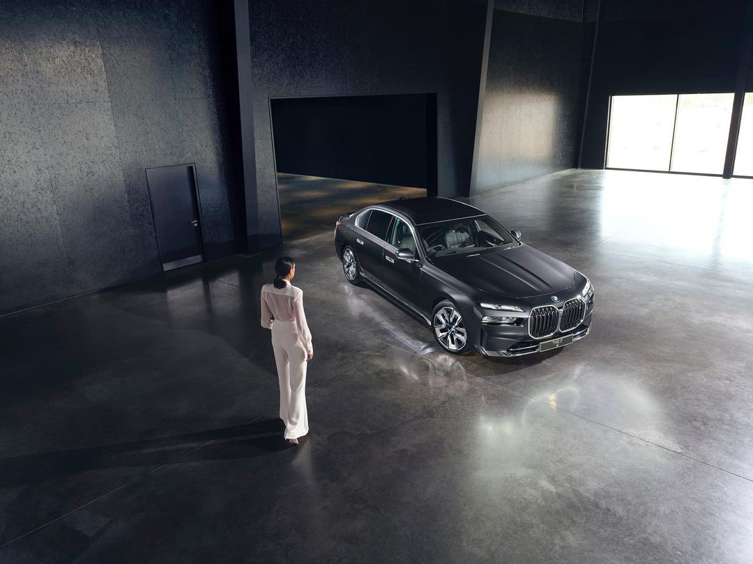 Woman in white stands near a modern black BMW 7 Series in a vast concrete room with large windows. Automotive luxury lifestyle.