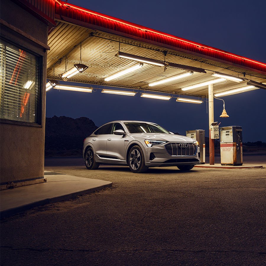 Silver Audi e-tron electric SUV parked at an old gas station at night with retro lighting and dark sky background.