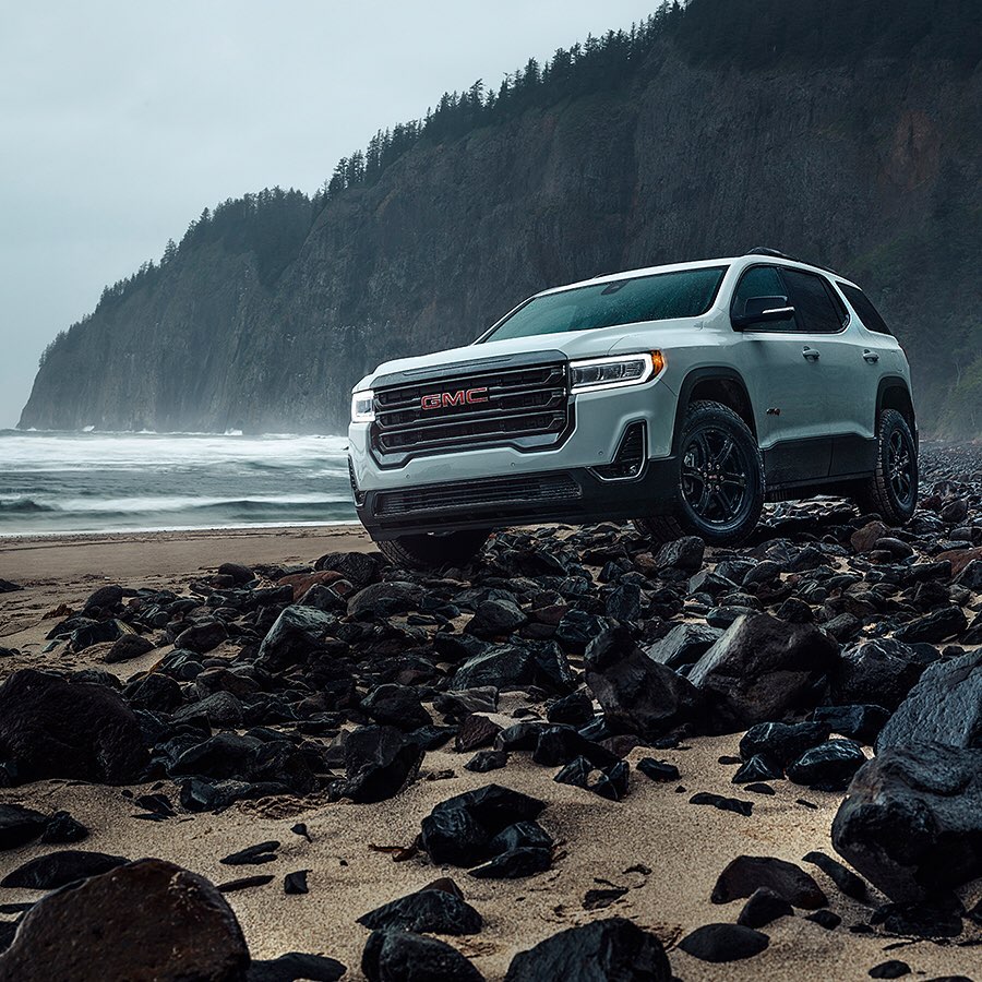 GMC Acadia SUV parked on a rocky beach with an overcast sky and a cliff in the background, showcasing automotive design.