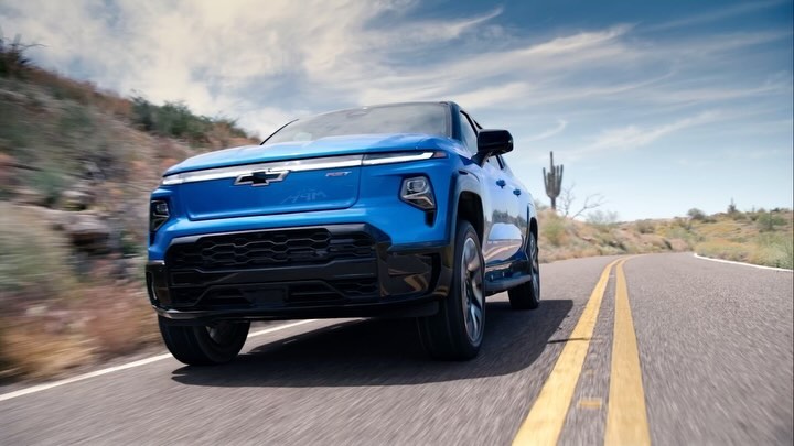 Bright blue Chevrolet Silverado EV driving on a desert road under a sunny sky with light clouds and arid scrub brush landscape.