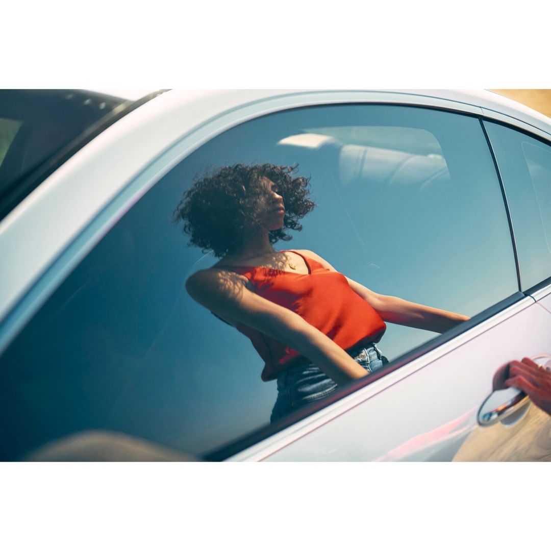 Young woman with curly hair is seen reflected in the window of a parked silver car on a bright day. The focus is on her reflection.