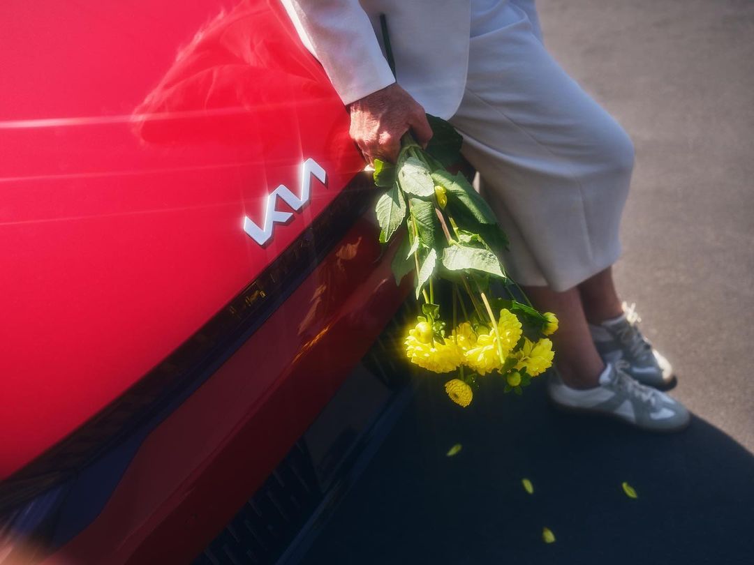 Red Kia car hood detail with person in white suit holding yellow flower bouquet, bright daytime lighting, and minimalistic composition.