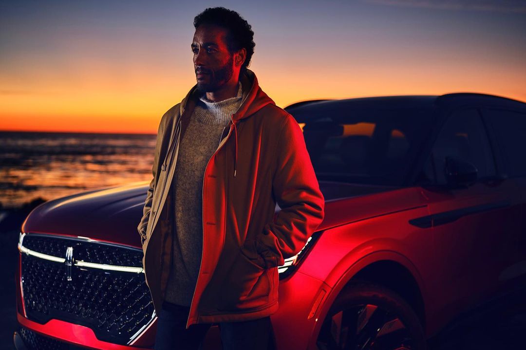Portrait of adult male in brown jacket near red SUV against sunset ocean backdrop, automotive fashion editorial, calm mood.