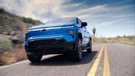 Bright blue electric pickup truck driving along a winding asphalt road through a desert landscape with sparse vegetation.