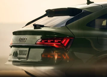 Rear three-quarter view of a dark green Audi luxury car parked outdoors during sunset with illuminated taillights.