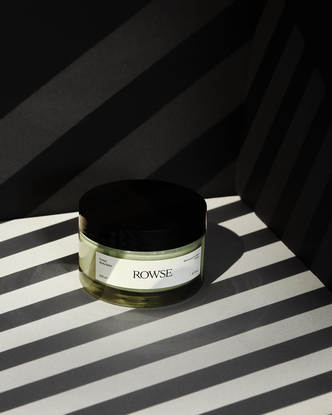 Glass jar of Rowse skincare product with black lid and white label on a surface with black and white stripes in a studio setting.