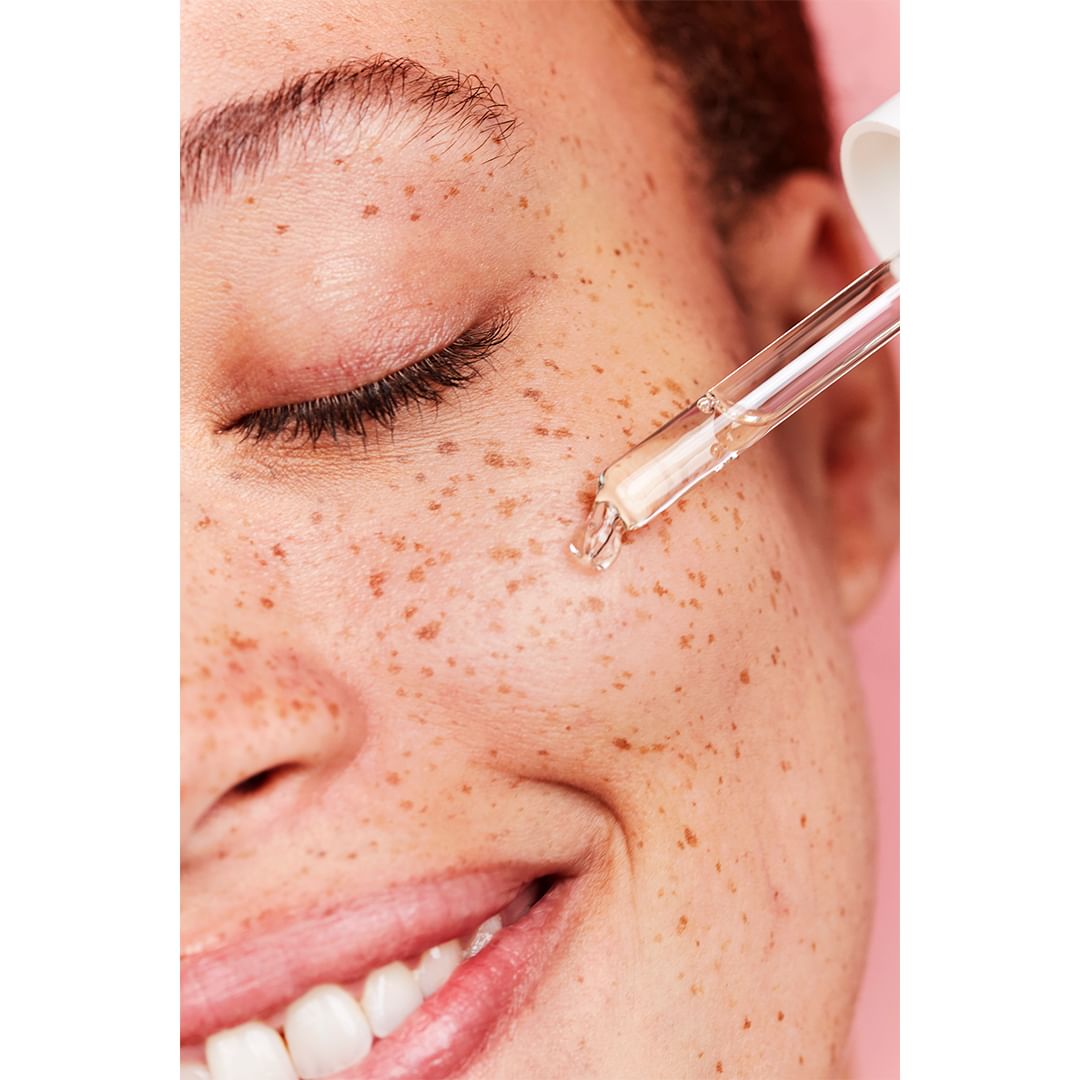 Close-up of a person's face with freckles, featuring an eyedropper applying serum to the skin against a pink background.