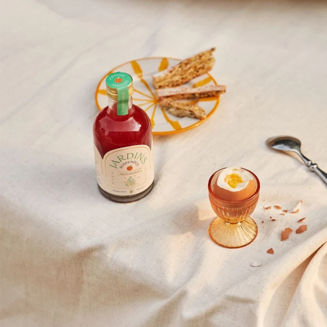 Still life of a bottle of red liquid, boiled egg in a glass holder, and biscotti on a plate, set on a white linen tablecloth.