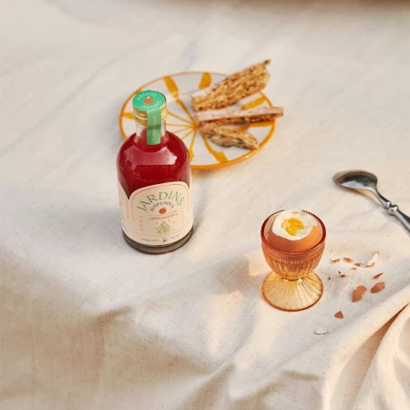 Still life of a bottle of red liquid, boiled egg in a glass holder, and biscotti on a plate, set on a white linen tablecloth.