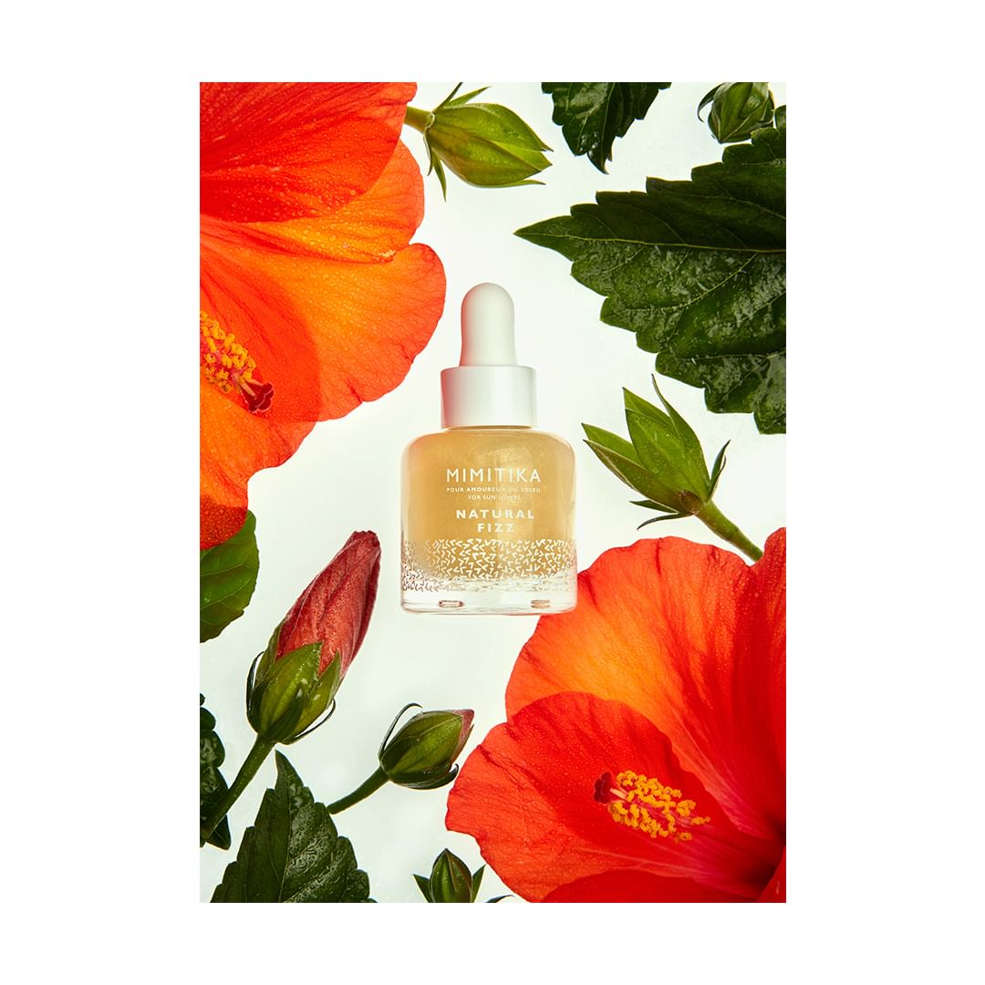 Mimikitka Natural Fizz bottle surrounded by red and orange hibiscus flowers and green leaves on a white background for beauty content.