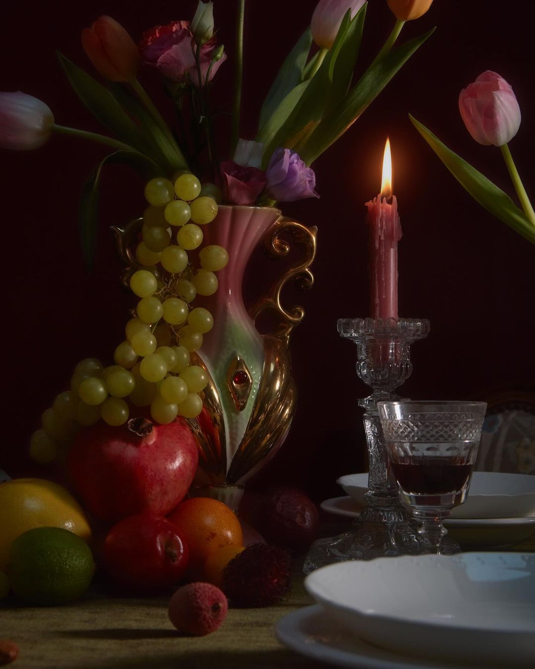 Still life photography: tulips, grapes, and mixed fruit in a golden vase, lit by candlelight on a table with white plates.