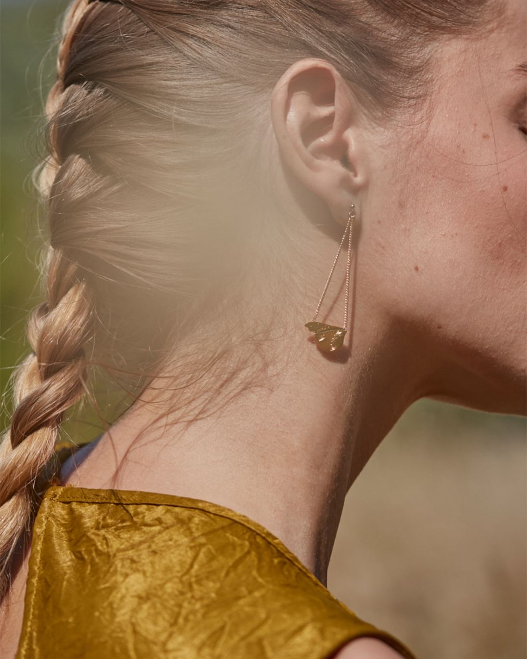 Adult woman with a blonde braid wearing a gold triangular earring and a gold textured dress, posing in a natural outdoor setting.