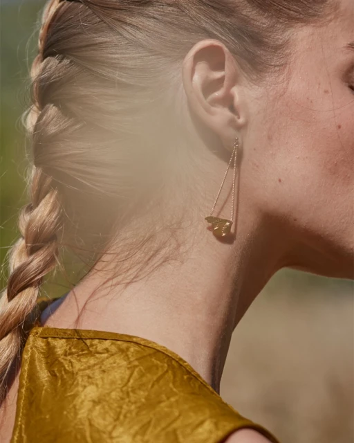 Adult woman with a blonde braid wearing a gold triangular earring and a gold textured dress, posing in a natural outdoor setting.