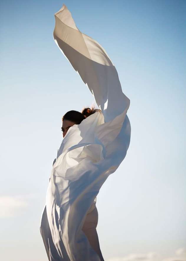 Adult female with wind-blown white fabric against a bright blue sky, creating a dynamic composition in natural light.