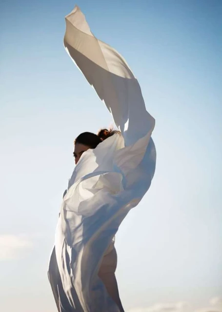 Adult female with wind-blown white fabric against a bright blue sky, creating a dynamic composition in natural light.
