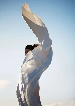 Adult female with wind-blown white fabric against a bright blue sky, creating a dynamic composition in natural light.