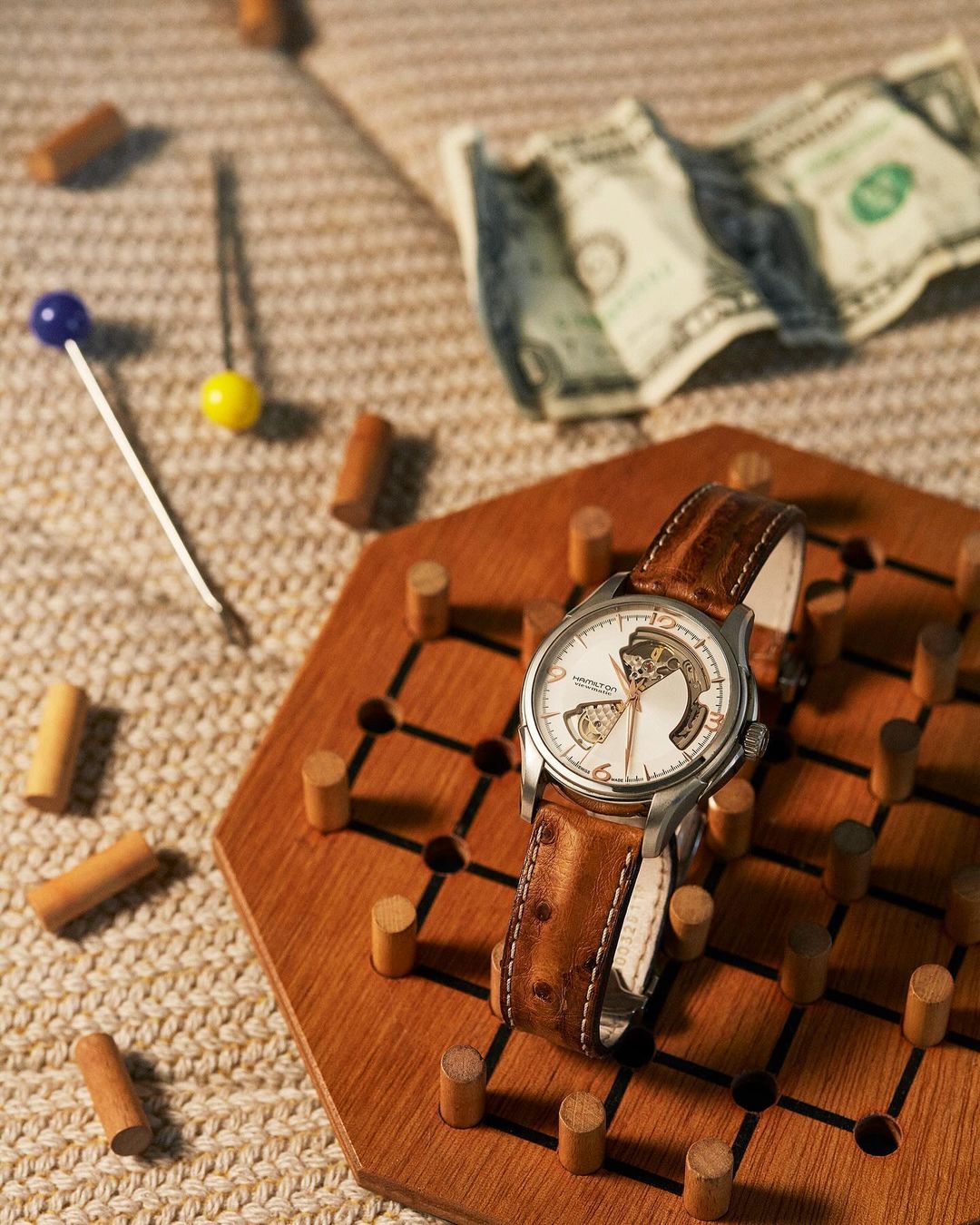 Luxury wristwatch with leather strap on wooden solitaire board with pegs, dice, and folded money on woven surface, product photography.