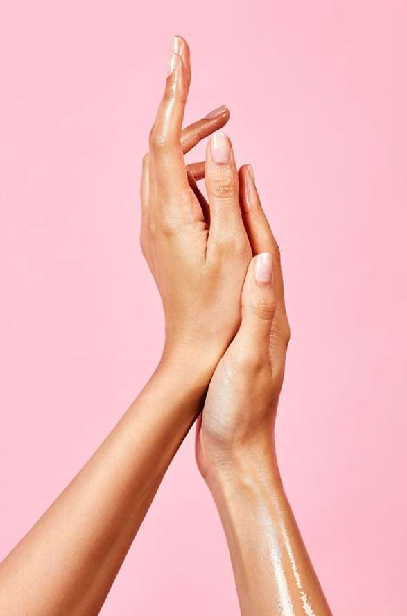 Close-up studio shot of a fair-skinned person's arms and hands covered in oil, palms facing each other, on a pink seamless backdrop.