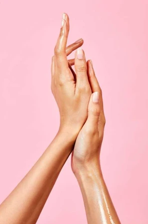 Close-up studio shot of a fair-skinned person's arms and hands covered in oil, palms facing each other, on a pink seamless backdrop.