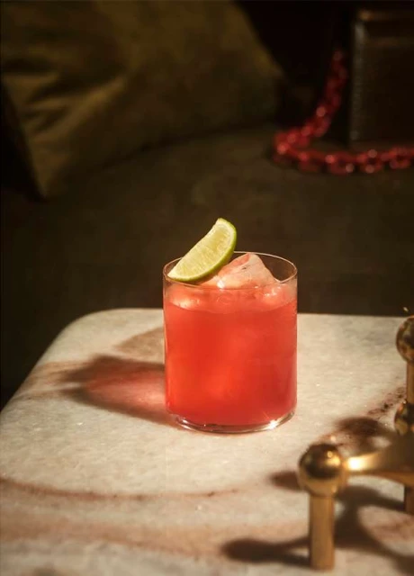 A refreshing red cocktail with ice and a lime wedge sits on a marble table, with a muted olive couch in soft-focus background.