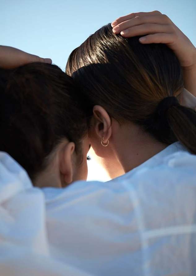 Close-up of two adult females embracing against a bright sky. They are wearing white shirts and gold earrings, hair up.
