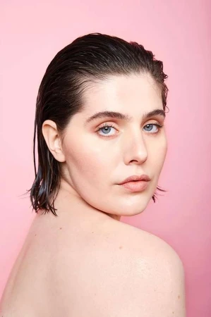 Close-up portrait of a woman with slicked-back wet hair and neutral makeup against a pale pink seamless backdrop.
