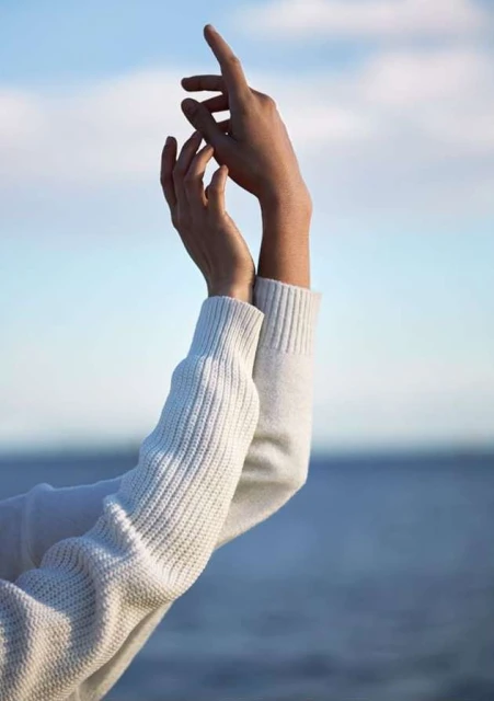 Close-up of hands reaching upwards against a sky and sea background, person wears a white knit sweater, neutral light.